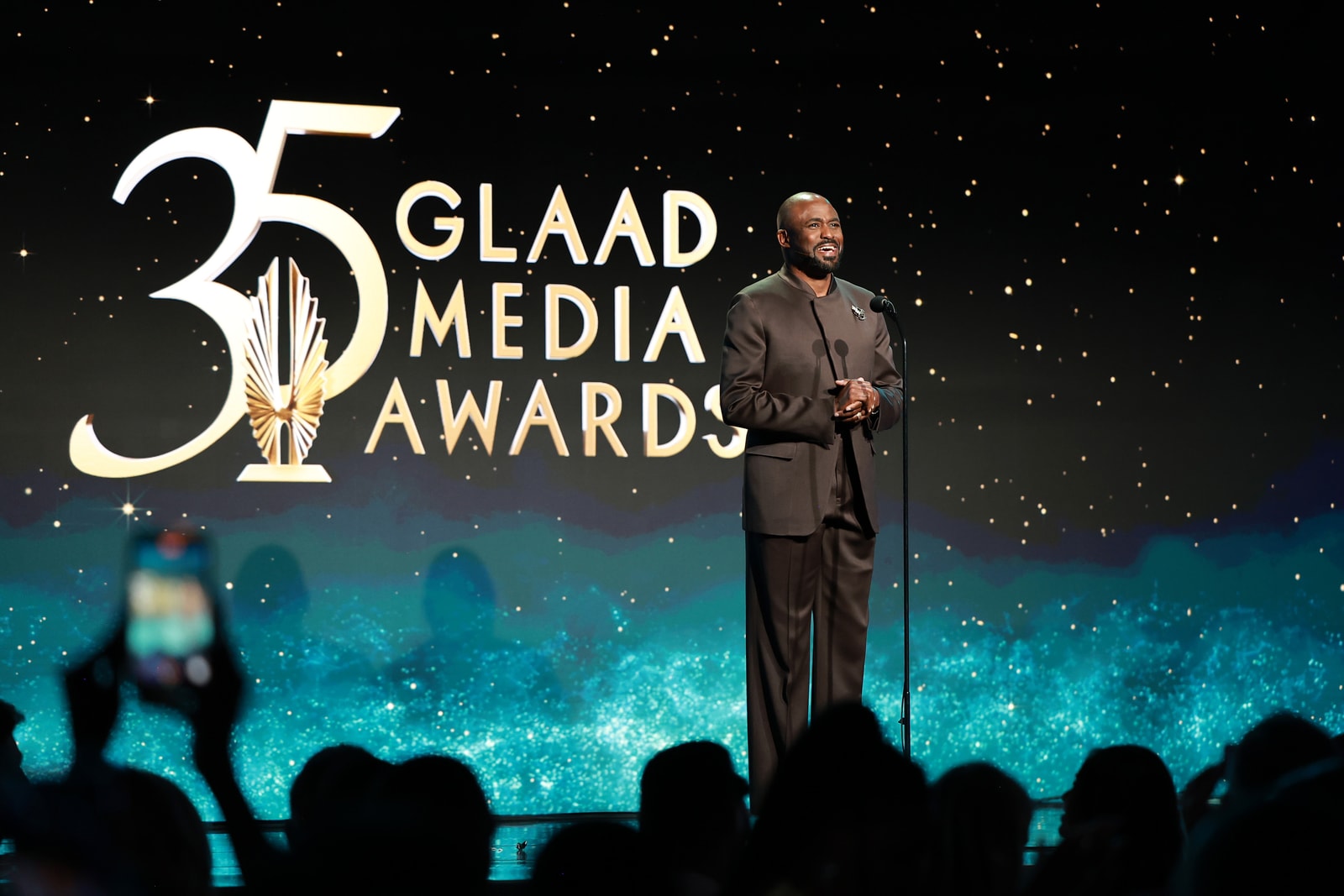 A speaker stands before the 35th GLAAD Media Awards logo and blue light effects.