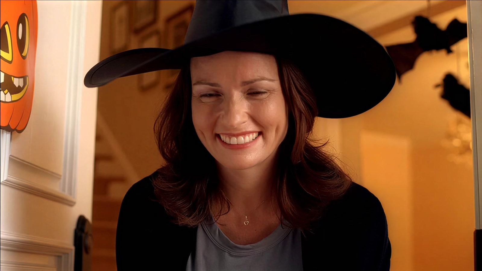 A smiling woman in a witch hat greets trick-or-treaters.